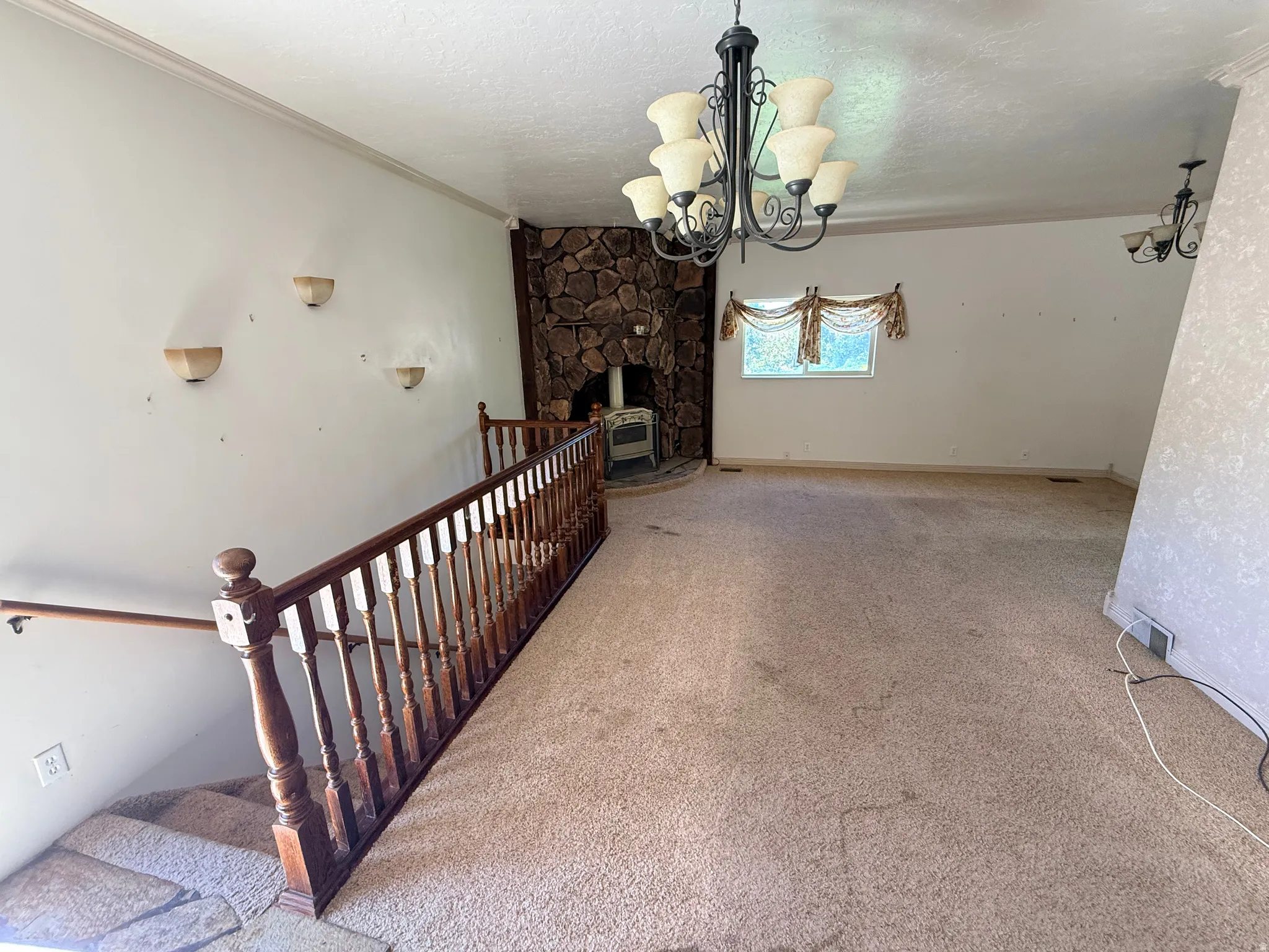 Spacious room featuring a stone-clad fireplace with a wood-burning stove, a window with decorative valance, a dark wood railing, and an overhead chandelier