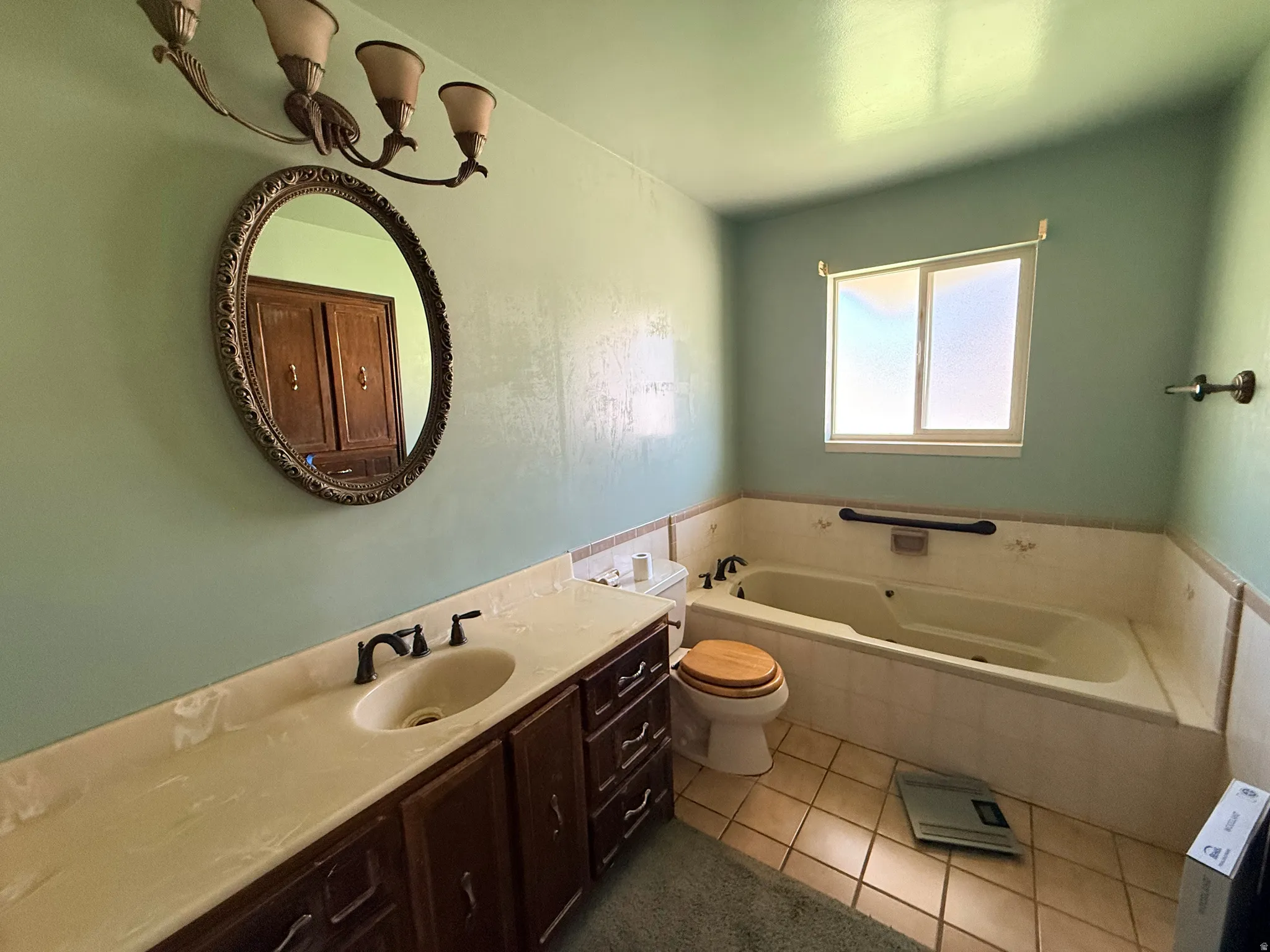 Soaking tub with tile surround, wood-finish vanity with an integrated sink, frosted window, and an oval vanity mirror with decorative frame