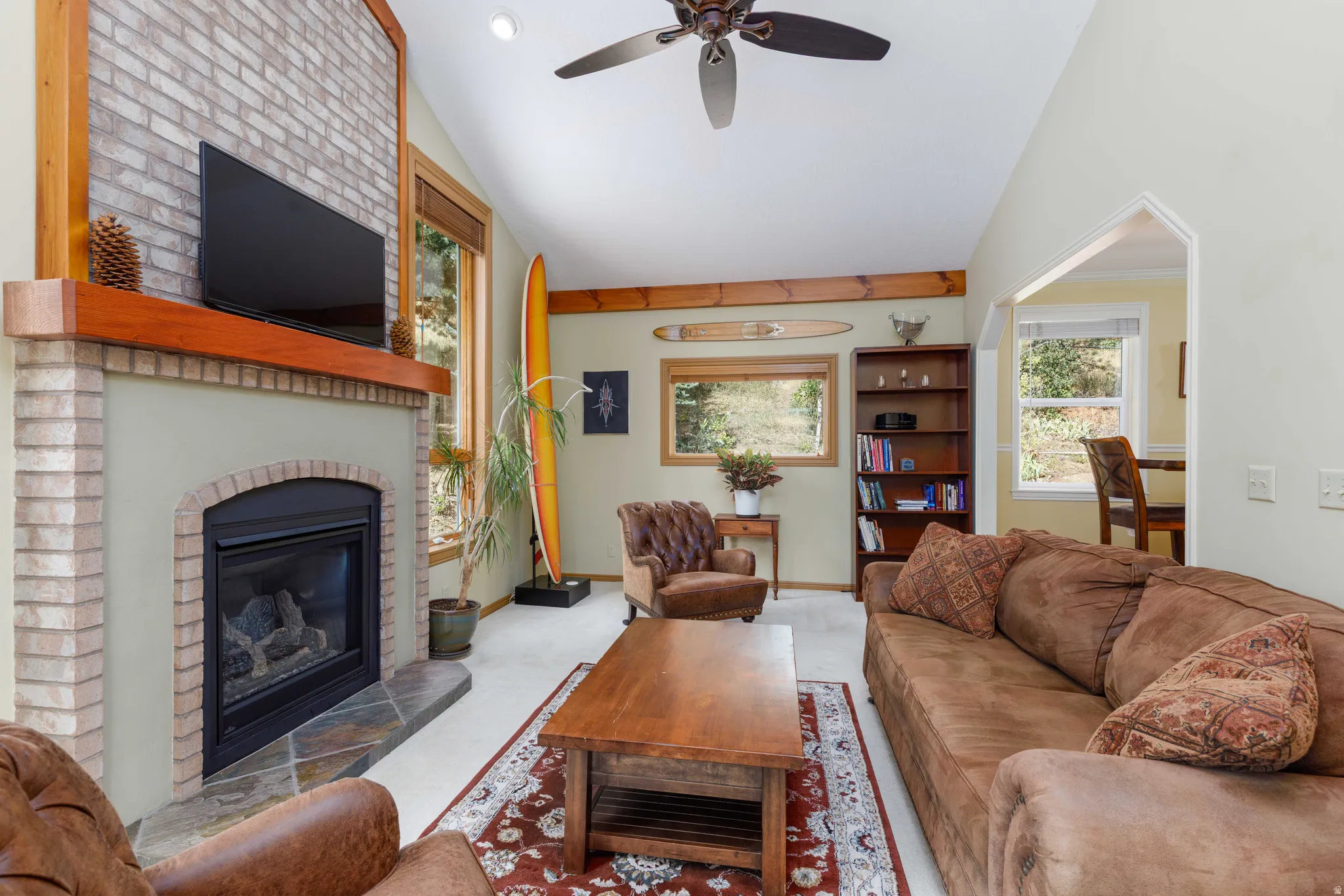 Vaulted ceiling with a ceiling fan, brick fireplace surround with a wood mantel, slate hearth, and wood-trimmed windows
