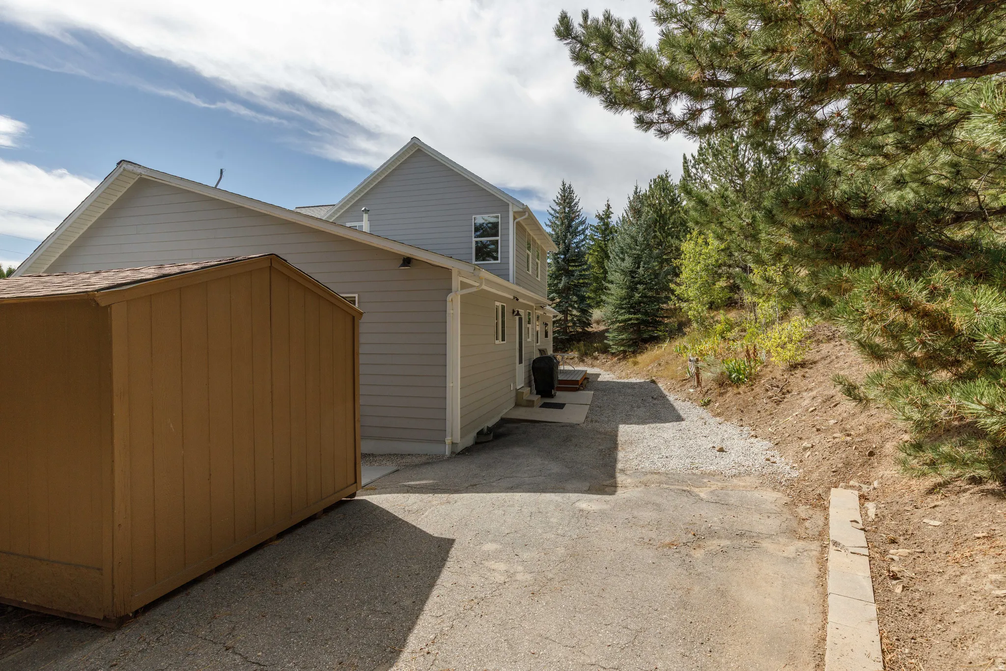 Wood-sided storage shed, gravel walkway, paved driveway, mature evergreen trees, and light gray siding