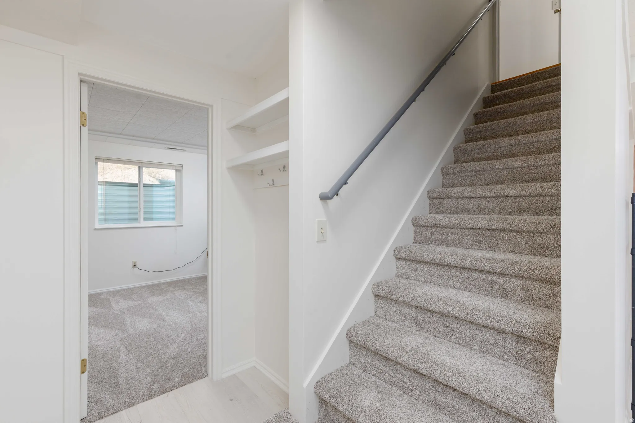Carpeted staircase with wall-mounted handrail, recessed wall shelving with coat hooks, and room with a window