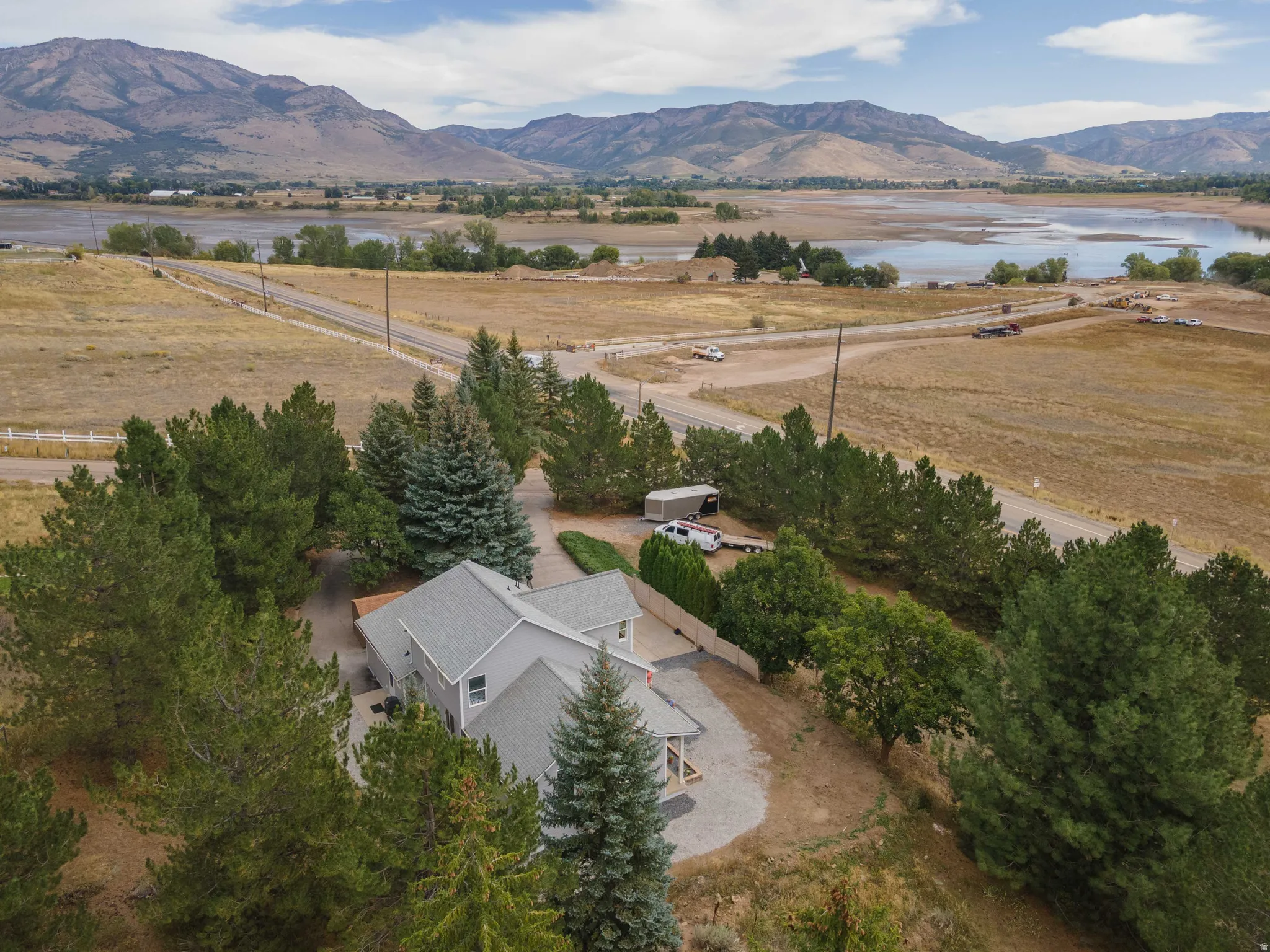 Gabled roofline, light gray exterior siding, mature evergreen trees, gravel driveway, and a wooden deck