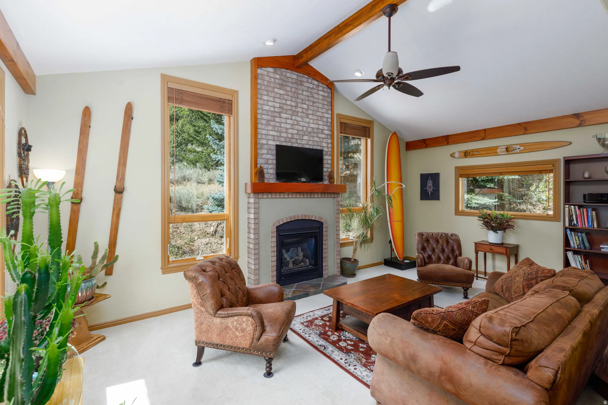 Vaulted ceiling with exposed wood beams, brick fireplace with a stone hearth, natural wood-framed windows, and built-in shelving