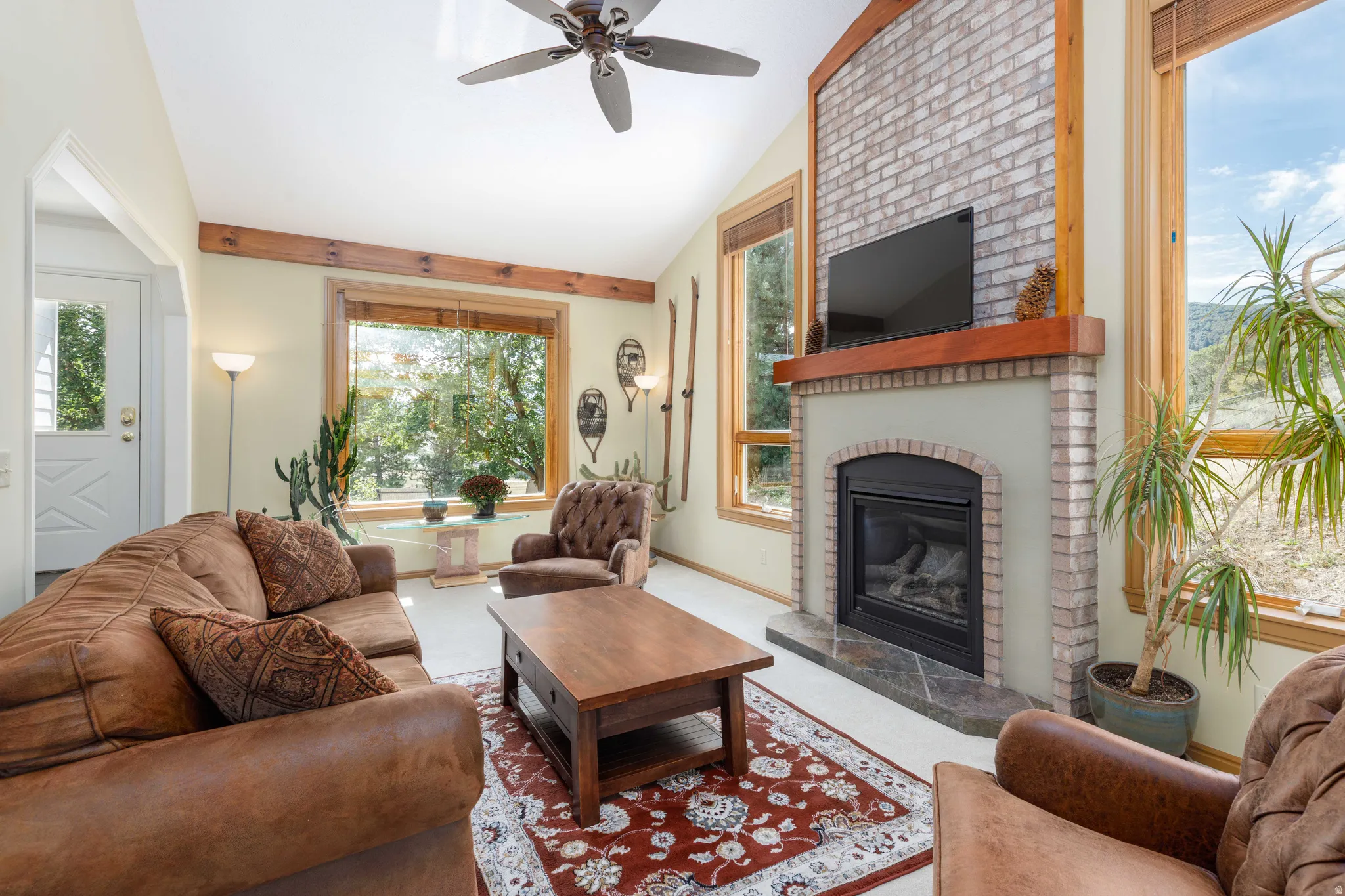 Vaulted ceiling, exposed wood beams, brick-surround fireplace with hearth, wood-framed windows, and a ceiling fan