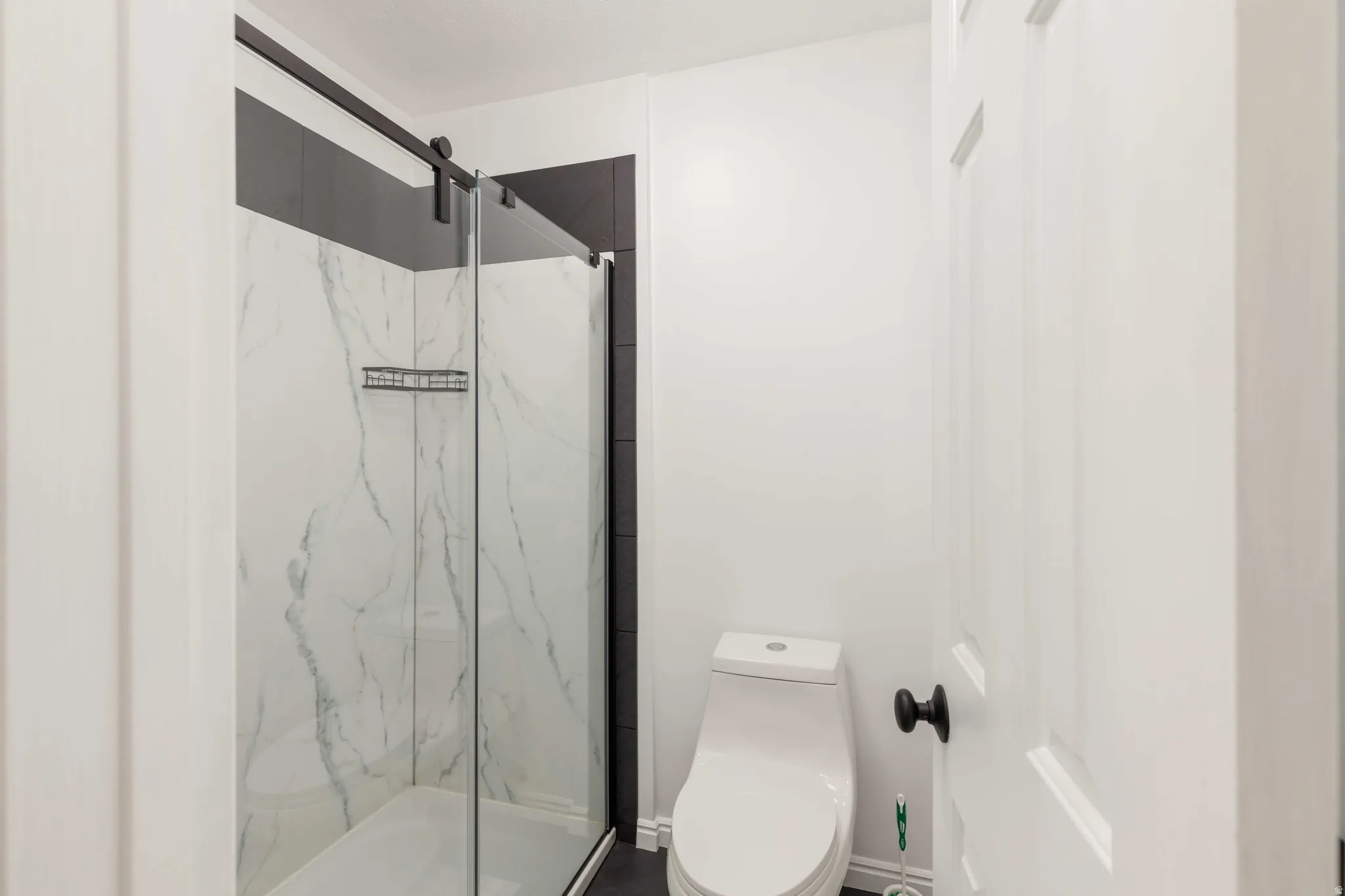 Glass-enclosed shower with a sliding barn door hardware system, marble-look paneling, and contrasting dark wall tiles
