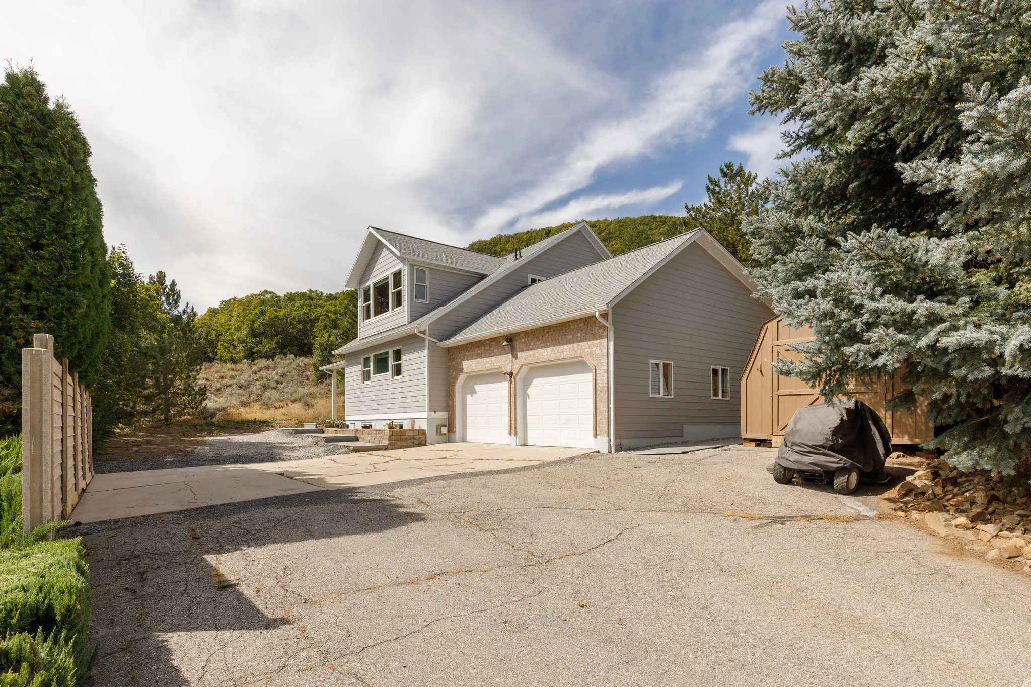 Two-car garage, light brick façade, gray siding, shed, and surrounding mature trees