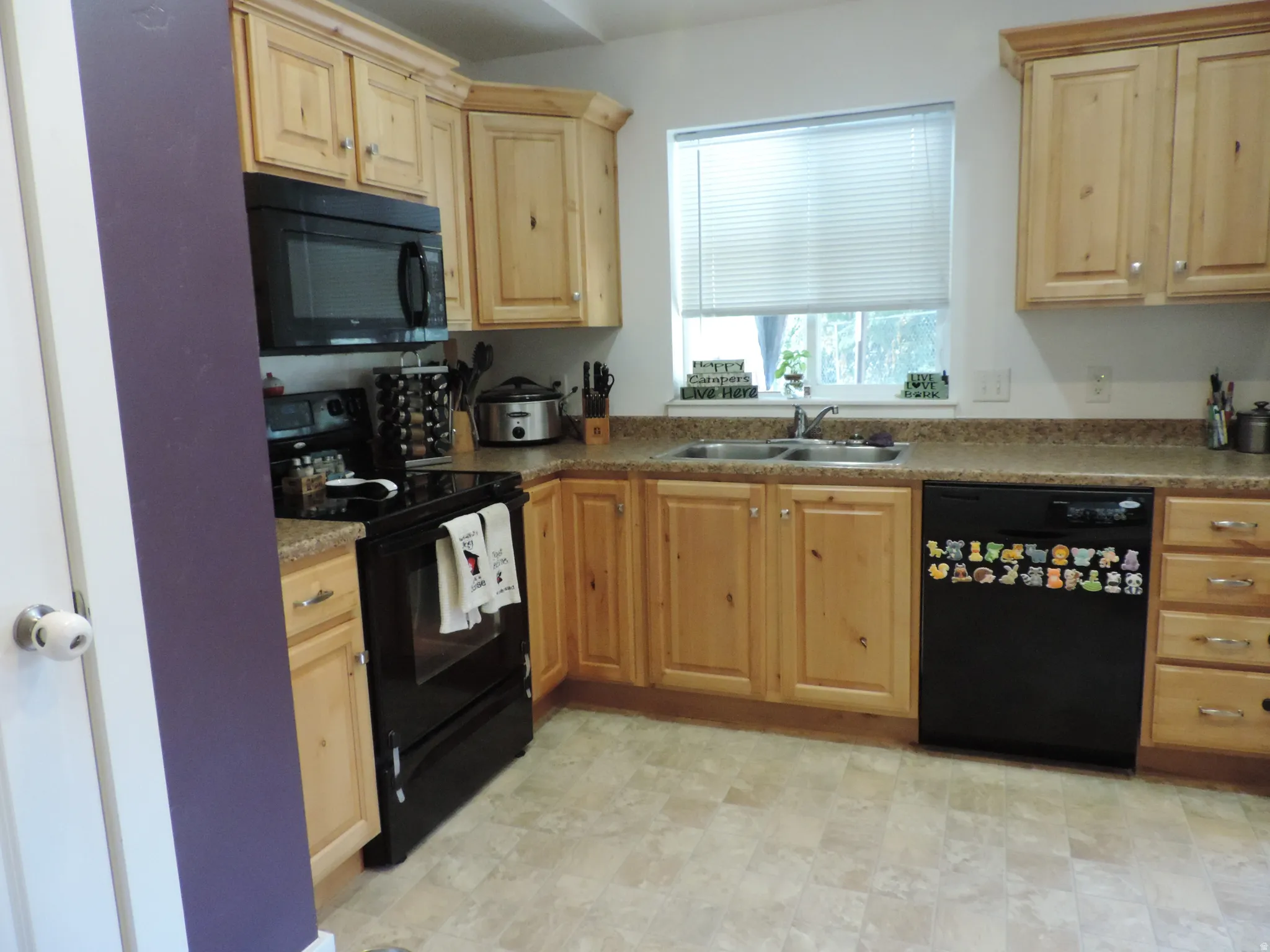 Kitchen featuring light wood cabinetry, black appliances, a double basin sink, and a window with blinds