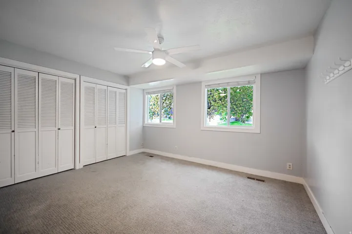 Recessed ceiling with integrated lighting, a ceiling fan, two windows, two sets of bi-fold louvered closet doors, and light-toned carpet flooring