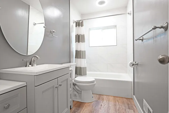 Wood-finish flooring, light grey vanity with integrated sink, oval mirror, white tiled tub surround, and a fixed window