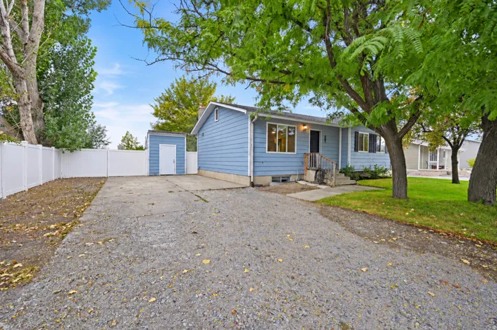 Light blue exterior siding, white window trim, mature trees, gravel driveway, and white privacy fencing