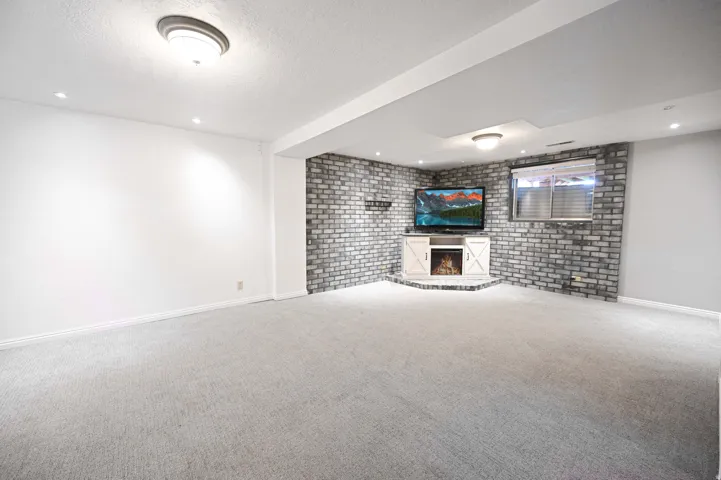 Spacious room with light gray carpeting, a corner accent wall with gray brick-finish, recessed lighting, and a white corner entertainment console with a built-in electric fireplace