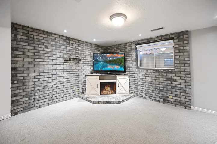 Textured brick accent wall, recessed lighting, built-in entertainment console with electric fireplace, window with horizontal blinds, and light-toned carpeting