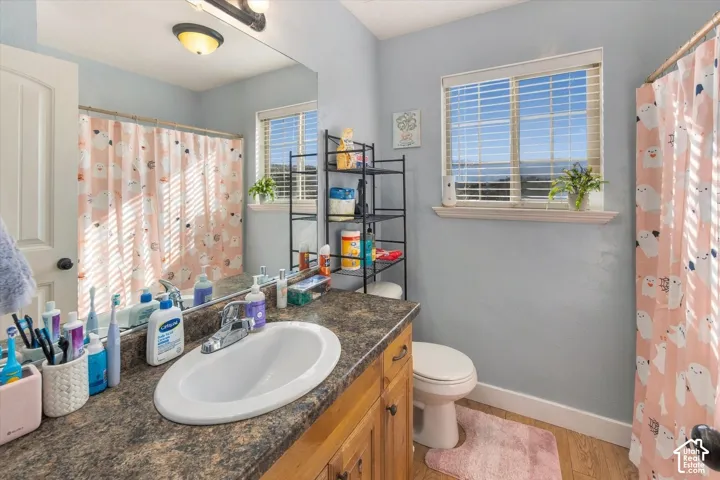 Bathroom featuring a wood-finish vanity with an integrated sink and chrome faucet, a large mirror, a window with blinds, and wood-finish flooring