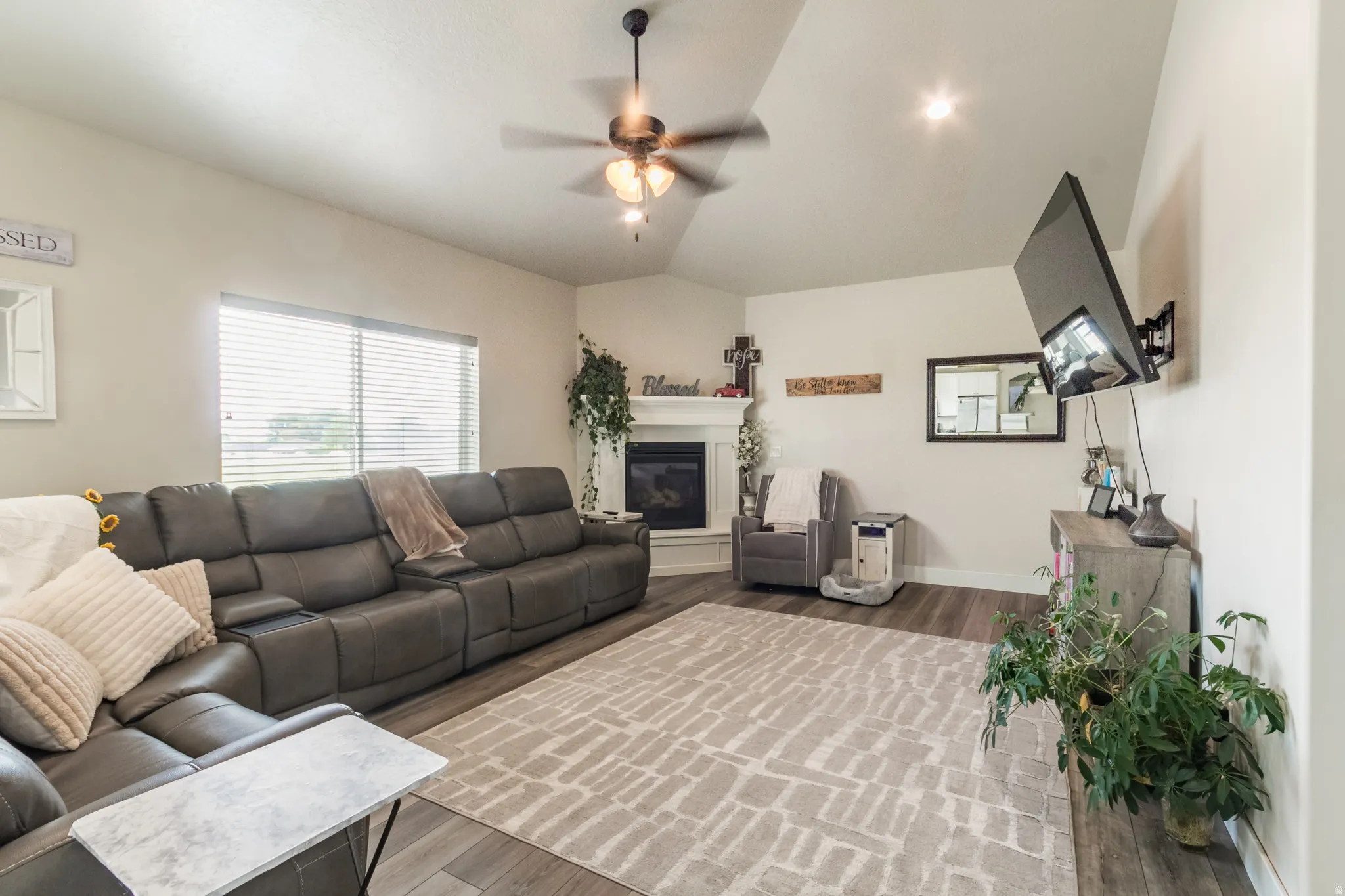 Vaulted ceiling with a ceiling fan and recessed lighting, wood-finish flooring, corner fireplace with a white mantel, and a large window with blinds