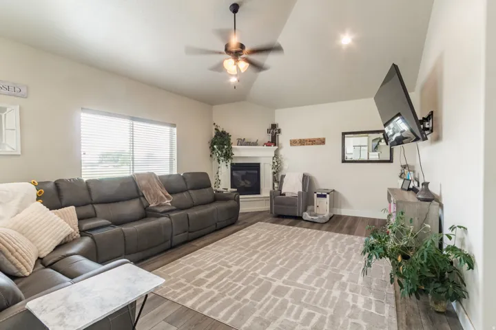 Vaulted ceiling with a ceiling fan and recessed lighting, wood-finish flooring, corner fireplace with a white mantel, and a large window with blinds