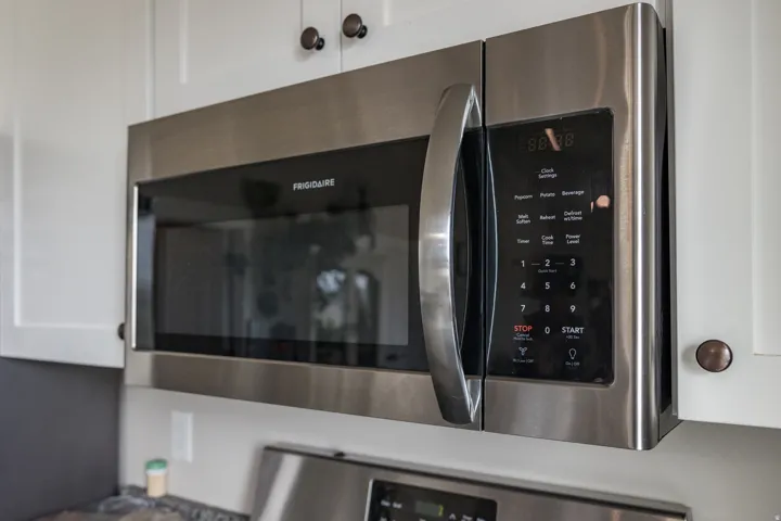 Stainless steel microwave oven with digital controls and a large handle, integrated into white upper cabinetry with dark bronze hardware