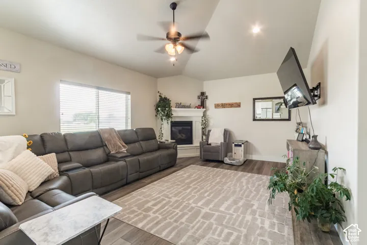 Vaulted ceiling with a ceiling fan and recessed lighting, wood-finish flooring, corner fireplace with a white mantel, and a large window with blinds