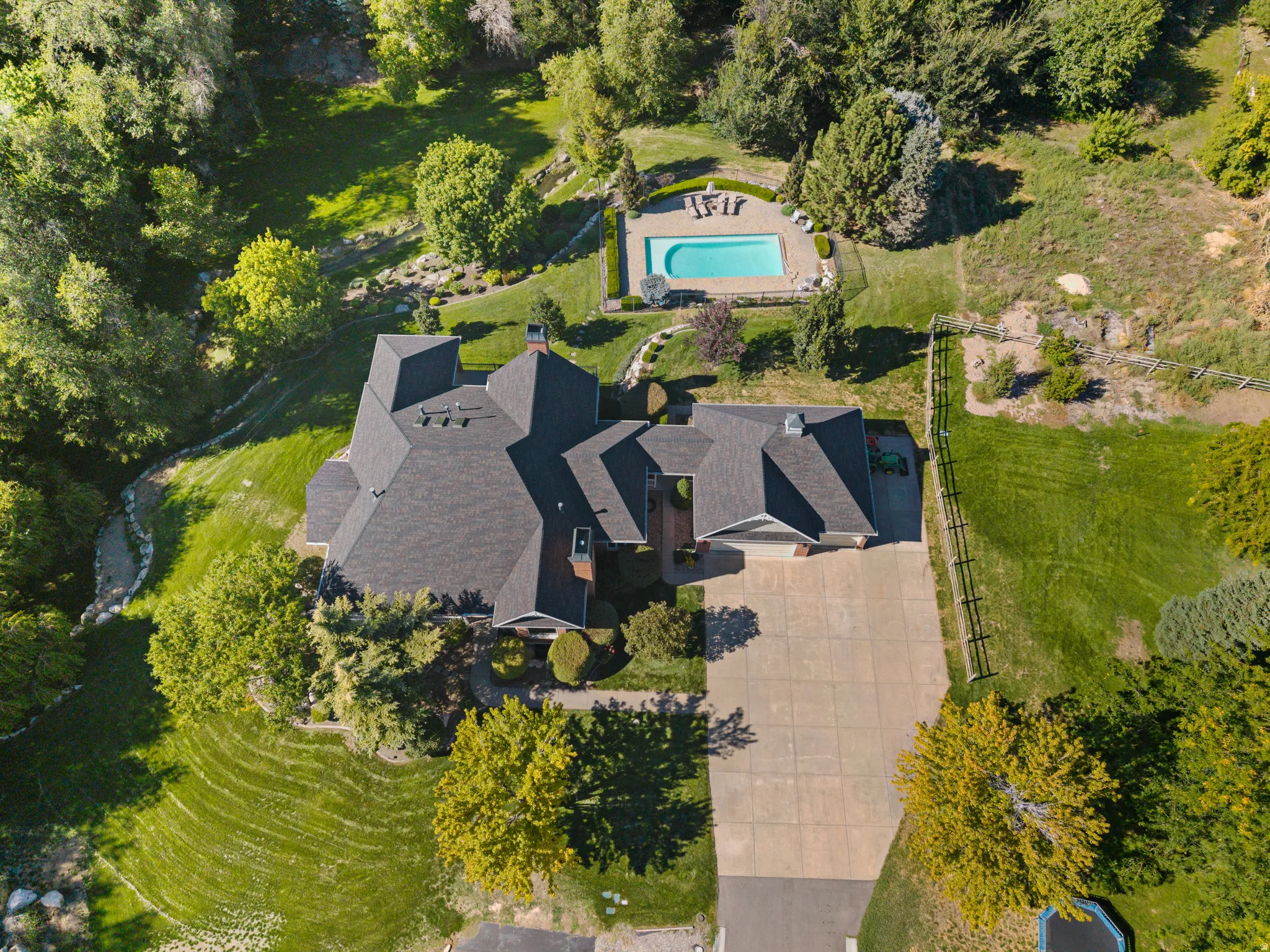 Expansive property featuring a multi-gabled roofline, concrete driveway, lush landscaping, and a rectangular swimming pool with paved surround