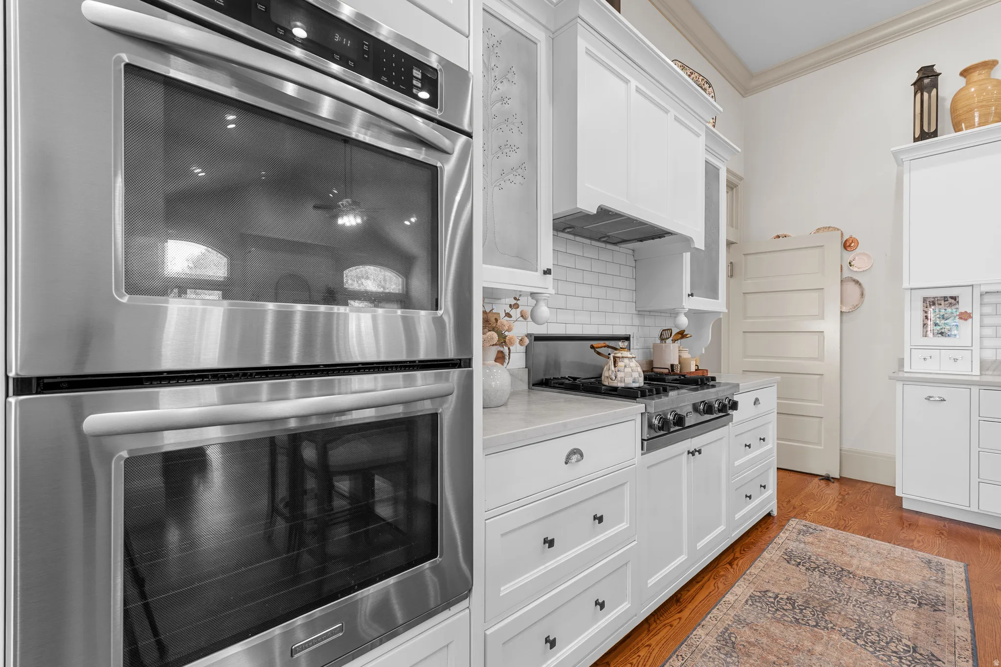 Stainless steel double wall ovens, gas range with hood, white shaker cabinetry, subway tile backsplash, and light-toned countertops