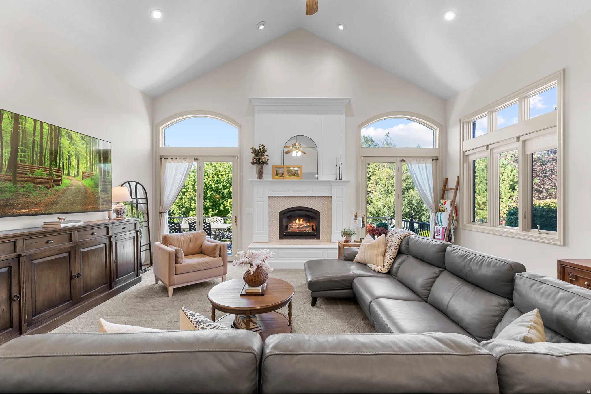 Vaulted ceiling with recessed lighting, gas fireplace with decorative mantel, multiple arched and rectangular windows, and glass panel doors opening to an outdoor patio