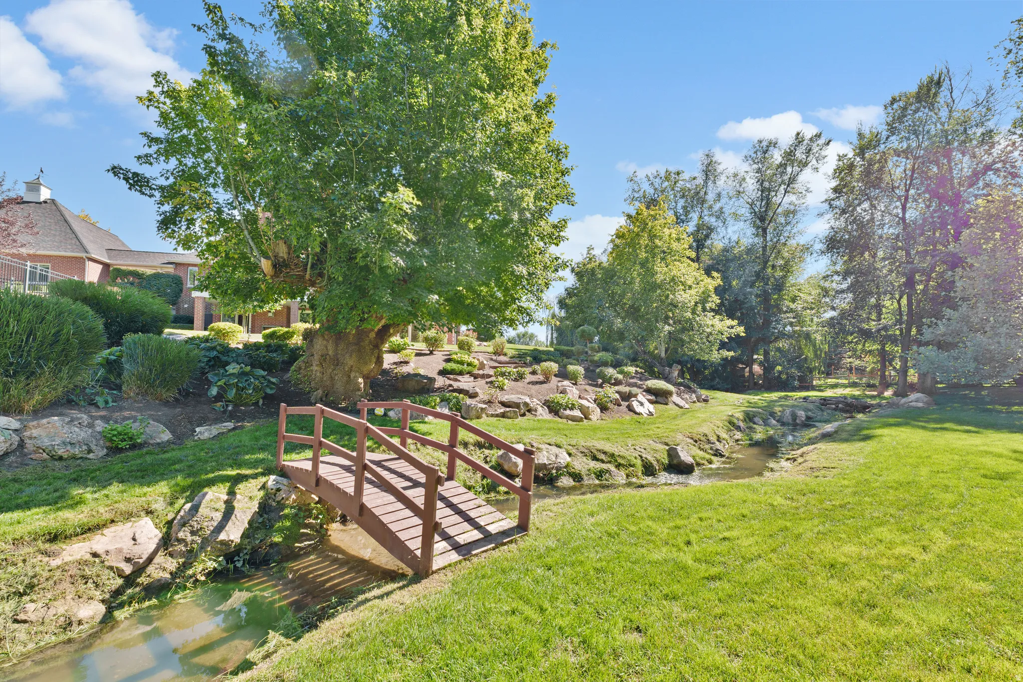 Wood-finish pedestrian bridge over a natural creek, surrounded by mature trees and landscaping