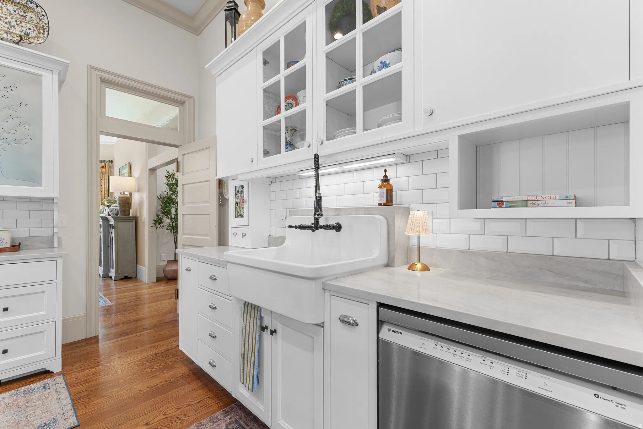 White cabinetry with glass-front upper doors, farmhouse-style sink with wall-mounted faucet, subway tile backsplash, and light-toned countertops