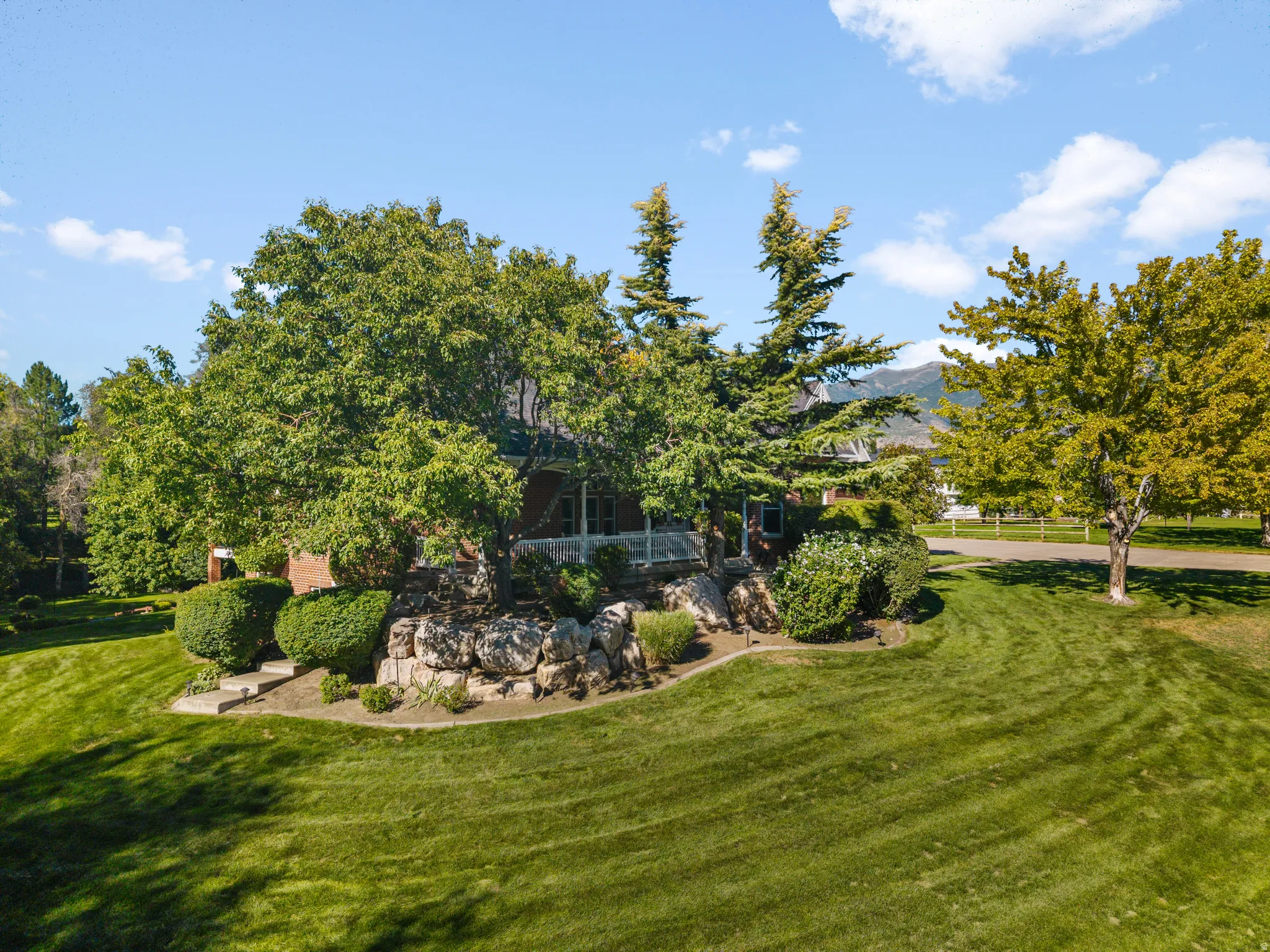 Expansive front lawn with mature trees, established landscaping, natural rock retaining walls, and a brick exterior