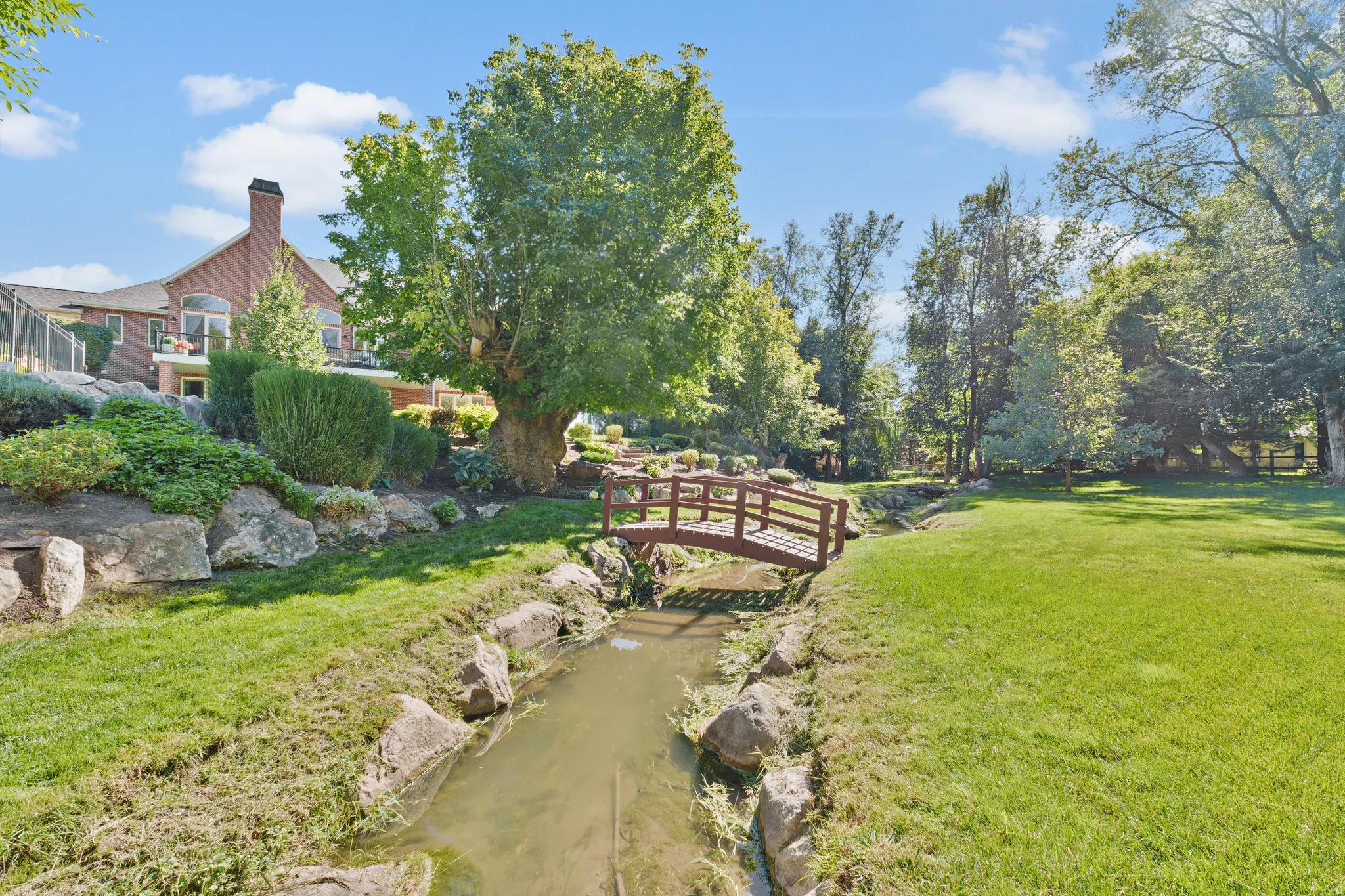 Expansive green space featuring a natural water feature, a decorative wooden bridge, mature trees, and established landscaping with natural rock elements