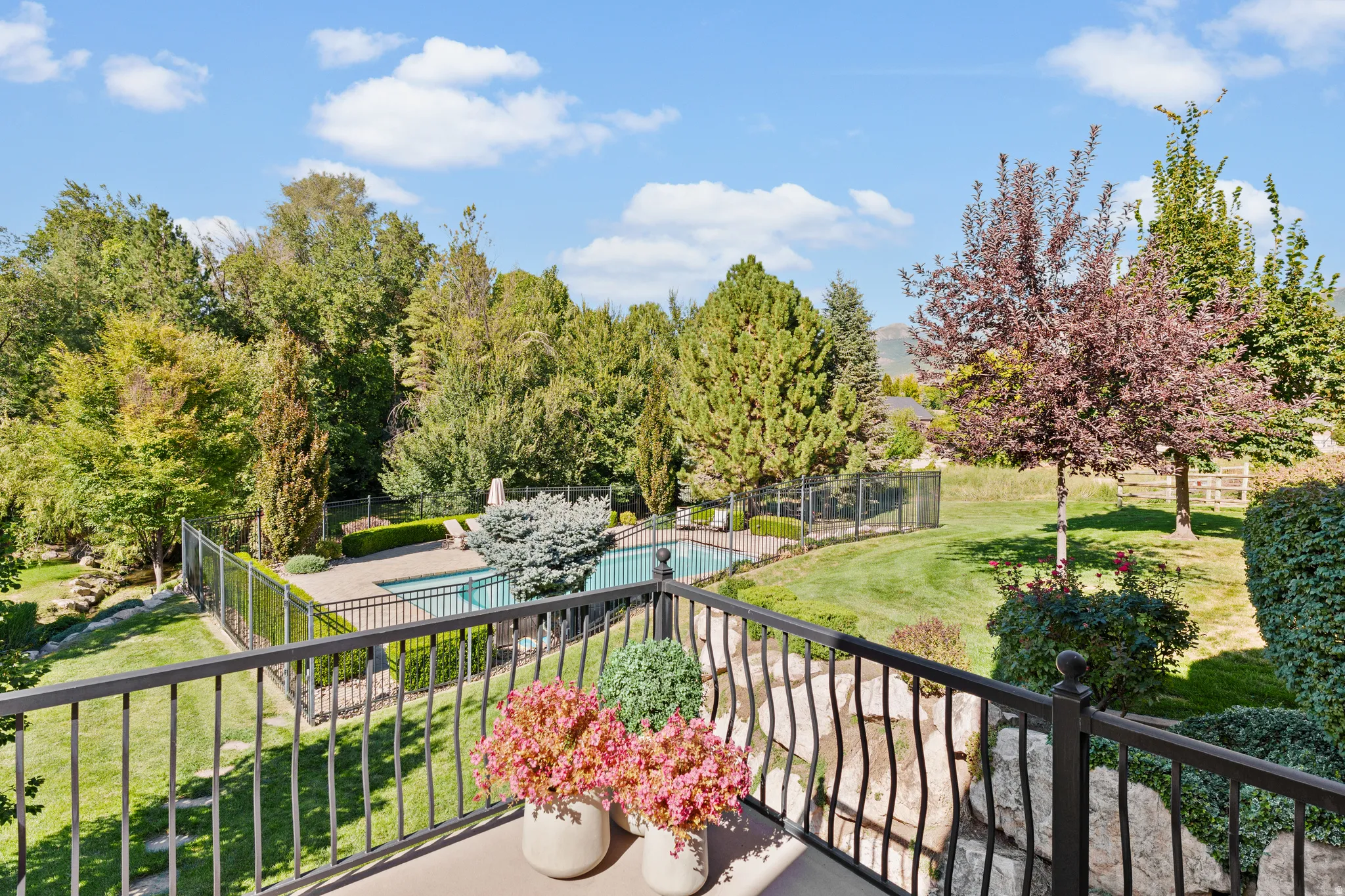 Outdoor swimming pool with surrounding patio, black metal fencing, manicured lawn, and mature trees