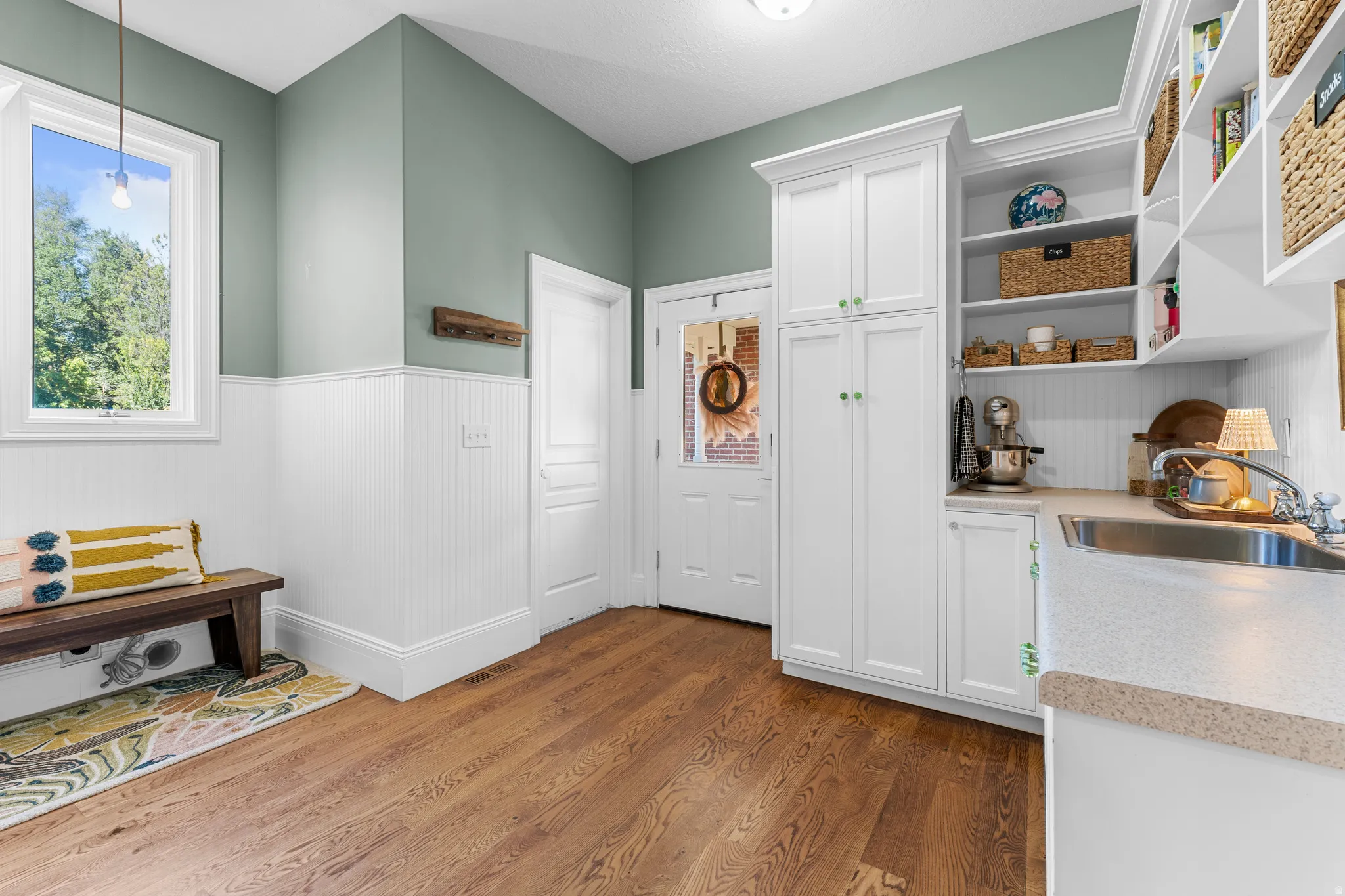 Wood-finish flooring, white beadboard wainscoting, white built-in cabinetry, stainless steel sink, and light-colored countertops