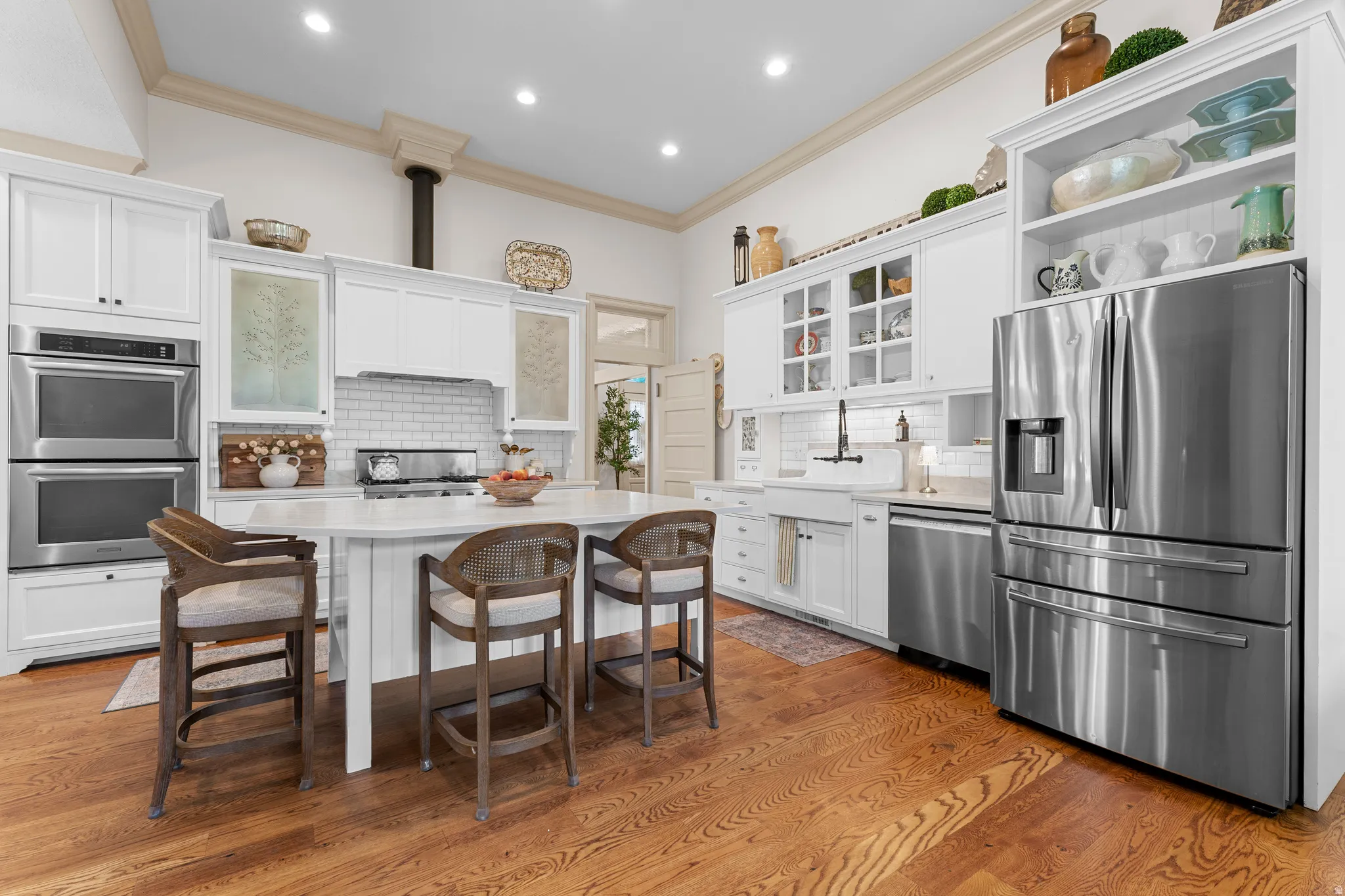 White cabinetry with integrated stainless steel appliances, a central island with a light-colored countertop, wood-finish flooring, a subway tile backsplash, and an apron-front sink