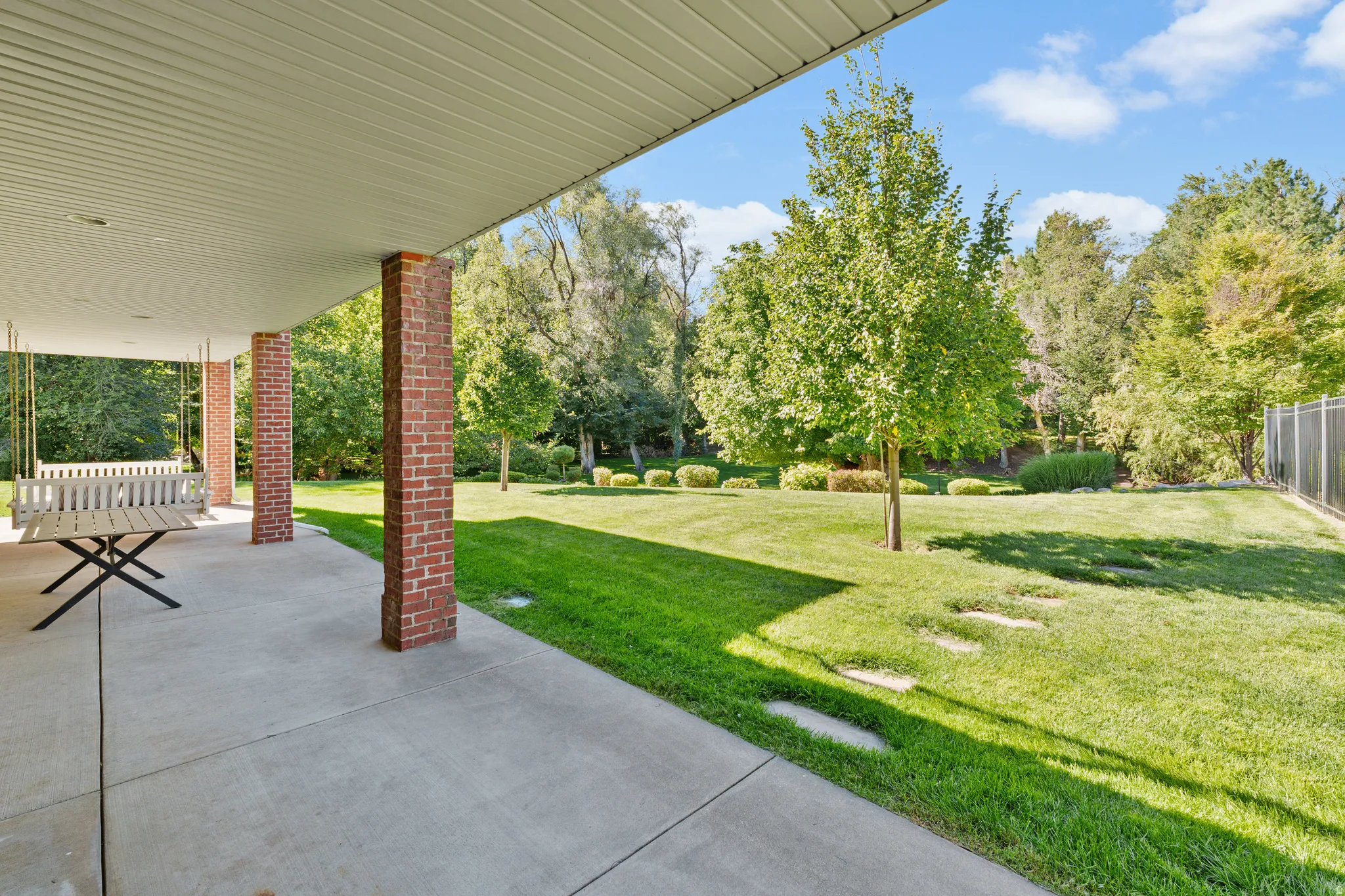 Expansive backyard featuring a manicured lawn, mature trees, and a covered concrete patio with brick support columns