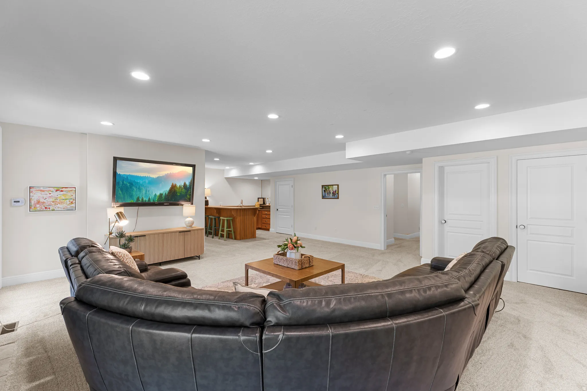 Recessed lighting illuminates this expansive space featuring neutral-toned carpeting, a built-in wood-finish bar, and multiple white panel doors