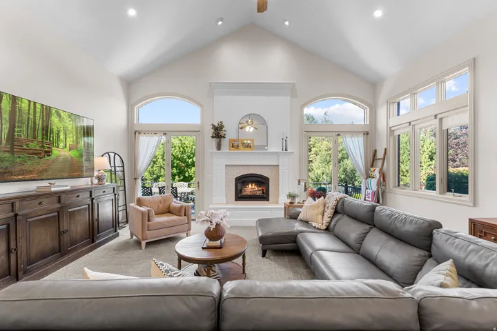 Vaulted ceiling with recessed lighting, gas fireplace with decorative mantel, multiple arched and rectangular windows, and glass panel doors opening to an outdoor patio