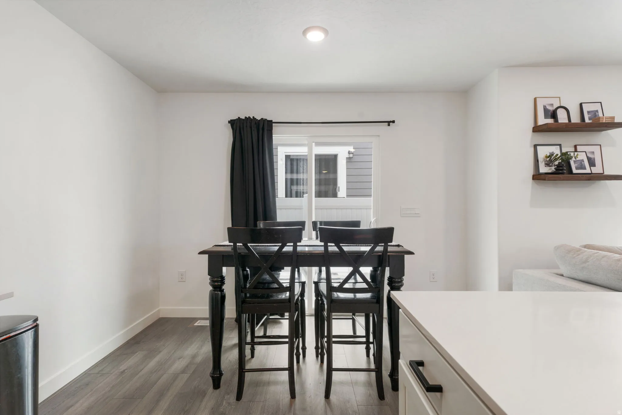 Bright dining area featuring a black wood table with matching cross-back chairs, large double-hung windows with black rod hardware and blackout curtains, recessed ceiling lighting, and dark wood-finish flooring throughout the open-concept space