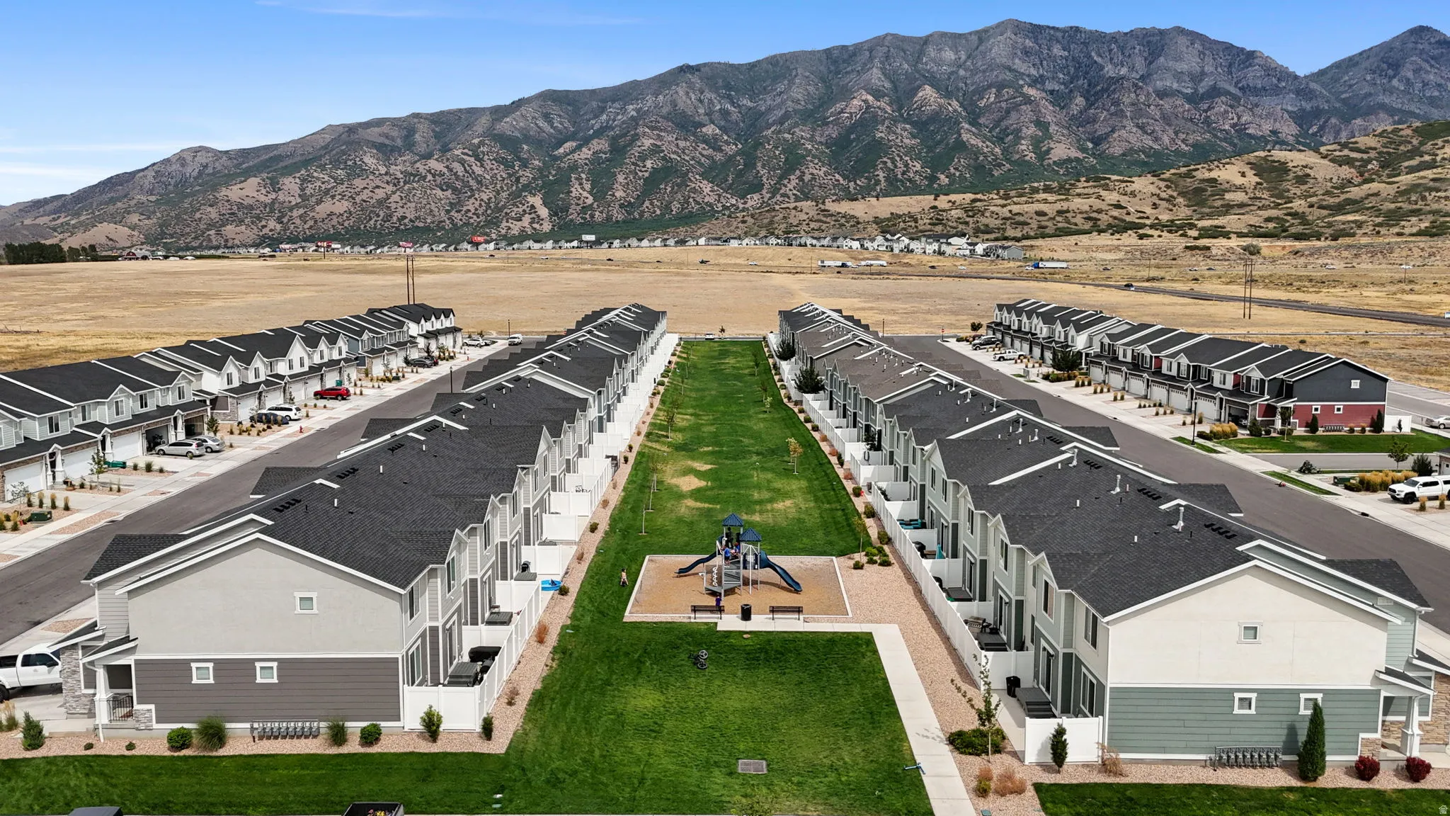 Modern townhome development featuring multiple residential buildings, a central green space with a playground, and mountain backdrops