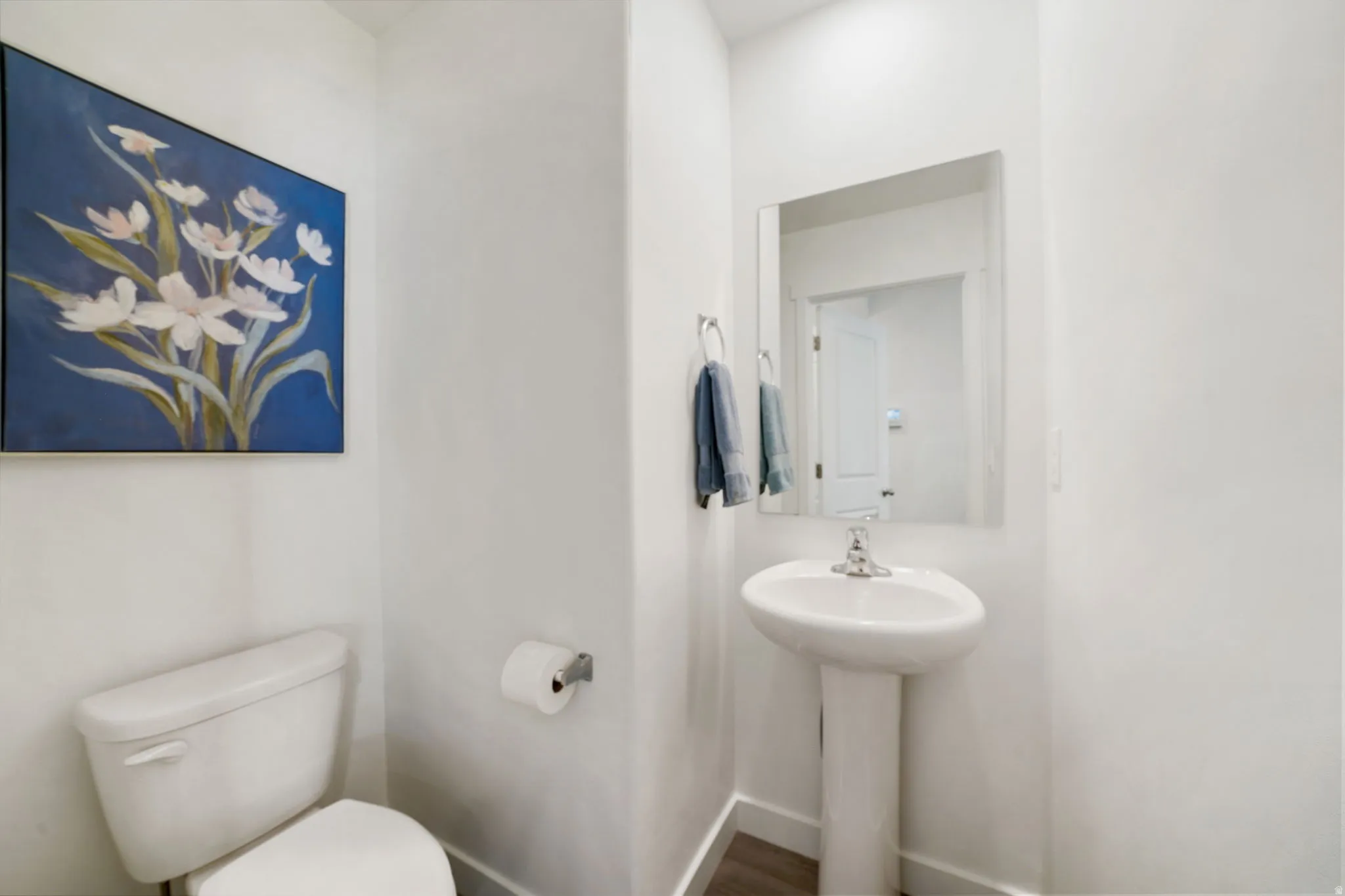 White pedestal sink, modern chrome faucet, wall-mounted mirror, and wood-finish flooring