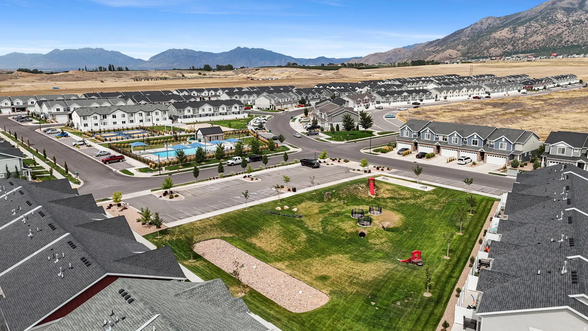 Community with a central amenity area, multiple residential buildings, paved streets, and mountainous terrain in the background