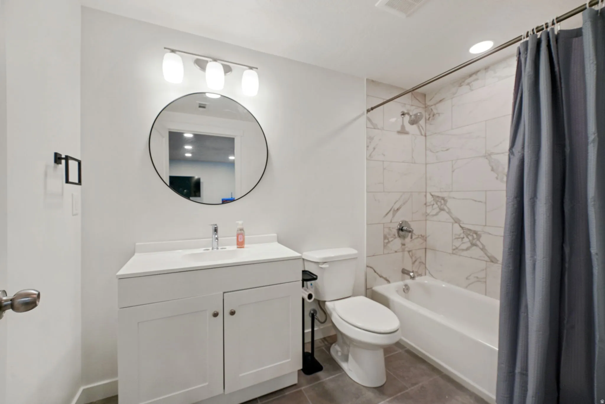 Modern bathroom featuring a white vanity with dual-door cabinetry and an integrated sink, a circular framed mirror with integrated lighting, a white toilet, and a bathtub/shower combination with marble-look tile surround and gray fabric shower curtain