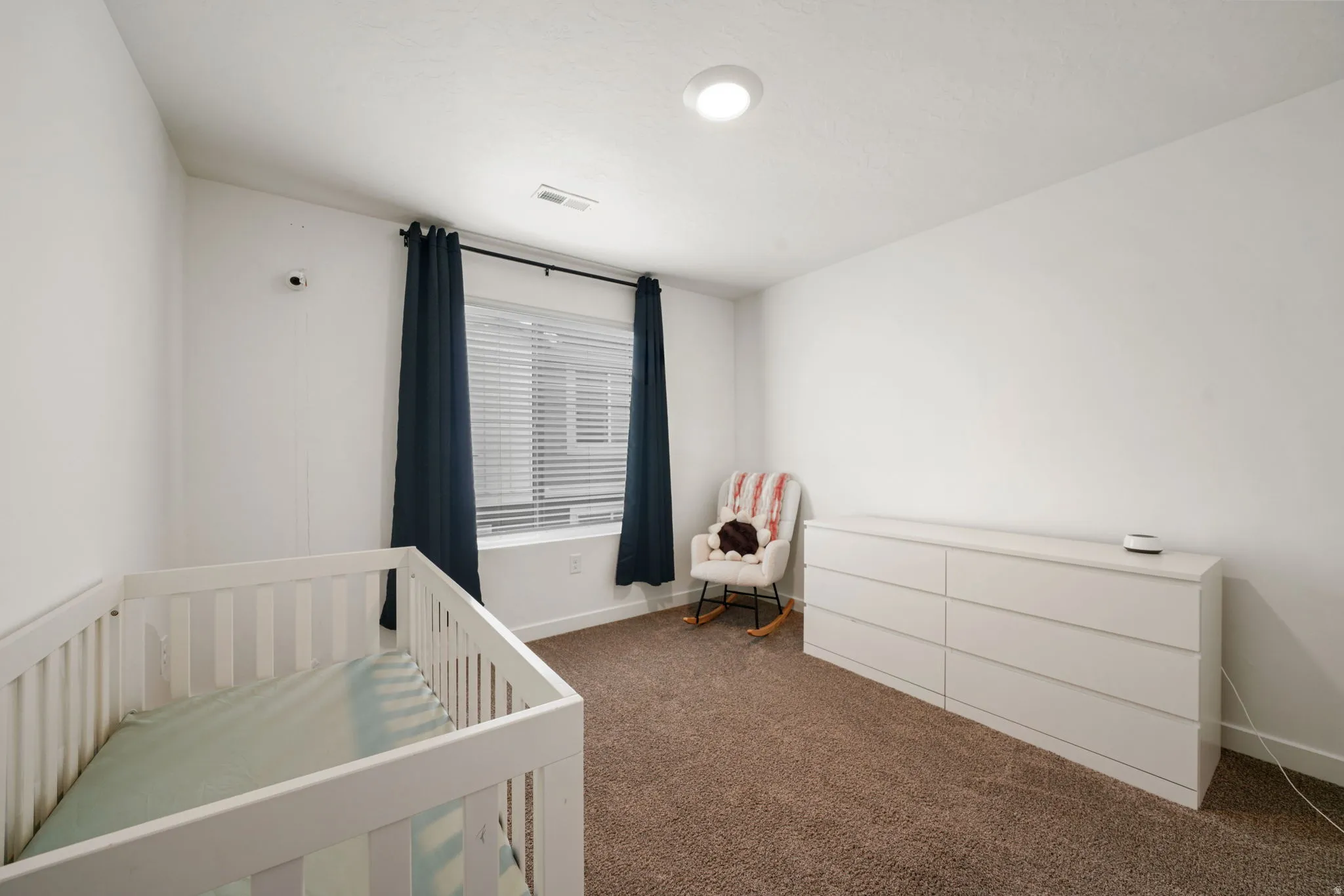 Bright nursery offering generous square footage with carpet flooring, a large window dressed in blackout curtains, white crib with coordinating changing table, and recessed ceiling lighting