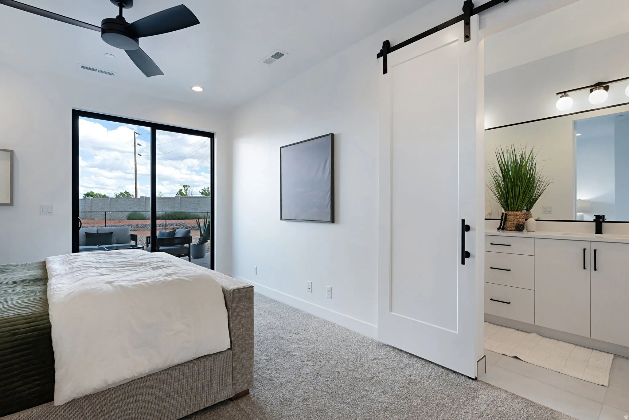 Black frame sliding glass doors, private outdoor patio, modern ceiling fan, white barn door with black hardware, and a double vanity with white cabinetry