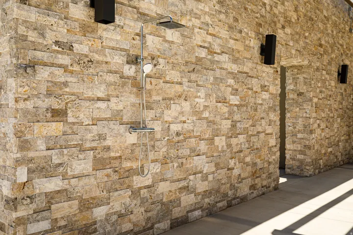 Textured stacked stone wall with a rain shower head and handheld shower, enhanced by two wall-mounted exterior light fixtures