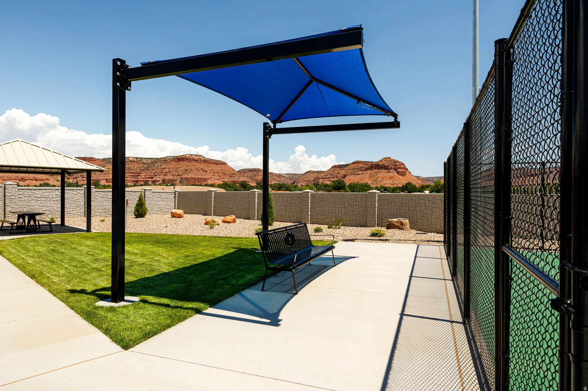Shaded outdoor seating area with a tensile fabric canopy, concrete pathways, manicured lawn, and a fenced court