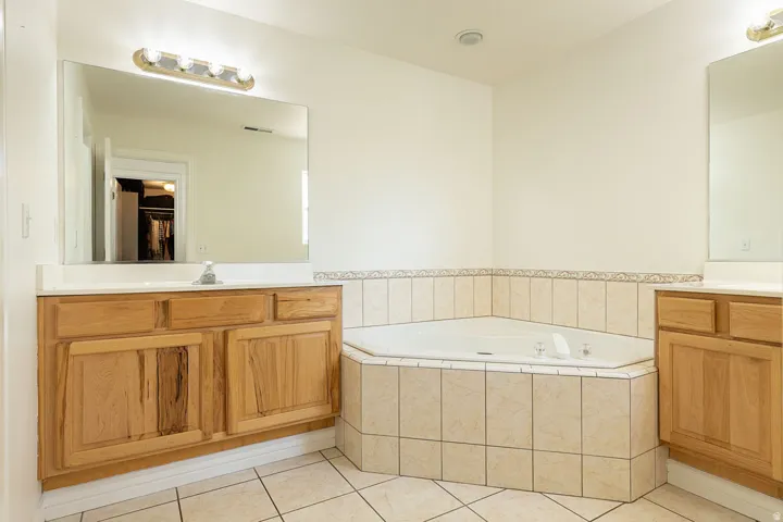 Dual wood-finish vanities with solid surface countertops, a corner soaking tub with tile surround, and tile flooring