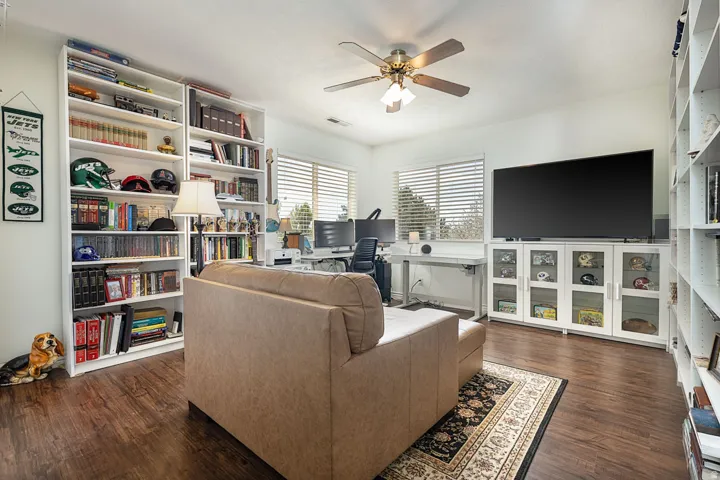 Dark wood-finish flooring with white built-in shelving, a ceiling fan with integrated lighting, and multiple windows with horizontal blinds