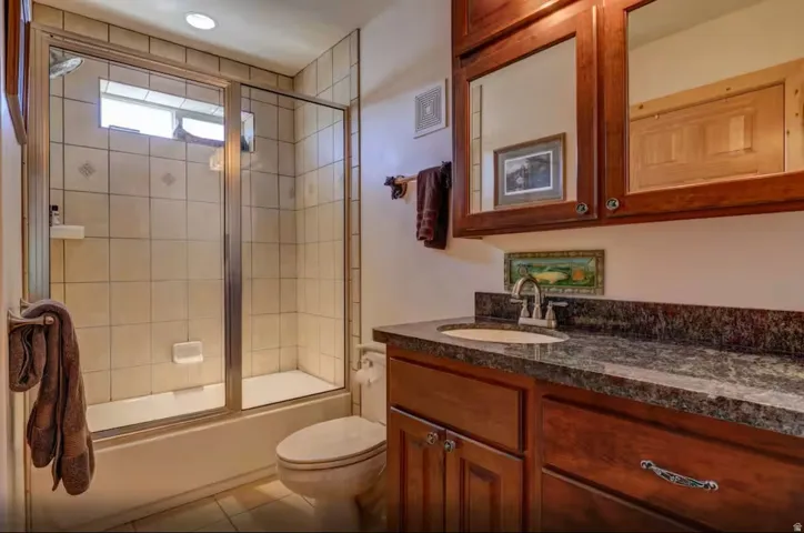Tiled shower with glass enclosure, wood-finish vanity with stone countertop, and upper wood-finish cabinetry