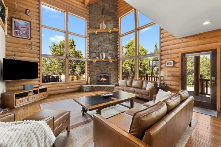 Vaulted ceiling with exposed wood beams, floor-to-ceiling windows, stone-clad fireplace, wood-finish flooring, and natural wood walls