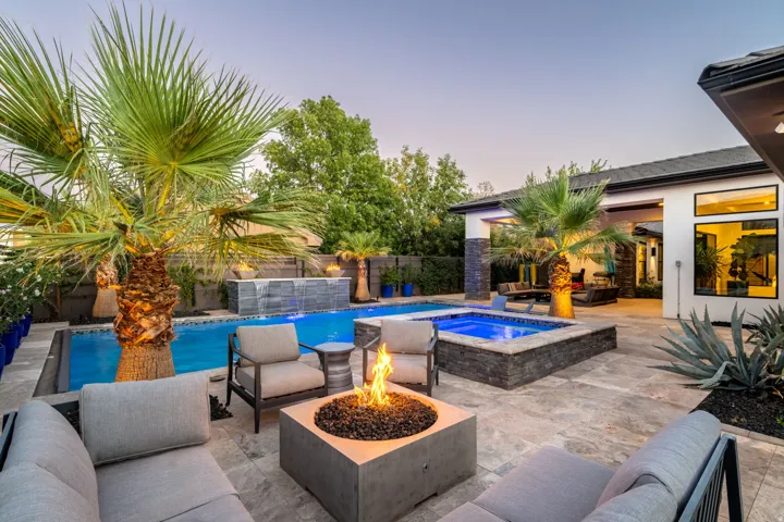 Expansive patio featuring a rectangular pool with an integrated spa, a tiered water feature, a fire pit, and mature palm trees