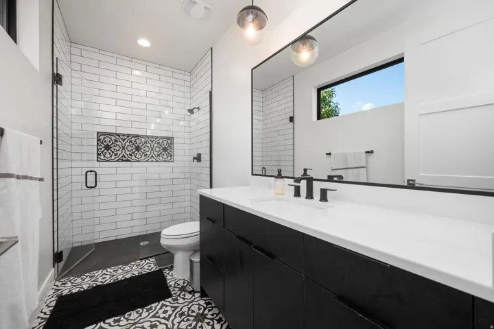 Black and white patterned tile flooring, a dark wood-finish vanity with a white countertop, and a shower with subway tile and glass enclosure