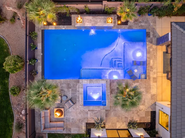 Expansive outdoor space featuring a rectangular swimming pool, integrated spa, multiple fire features, mature palm trees, and stone-tiled decking